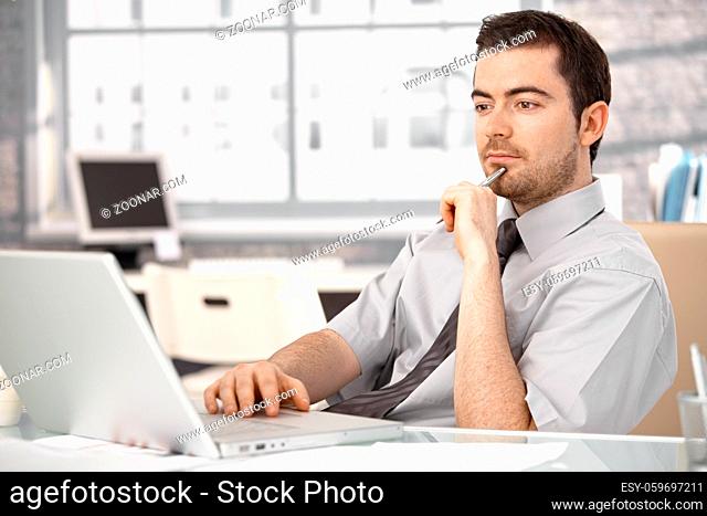 Young businessman working in bright office, sitting at desk, using laptop