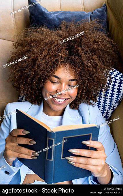 Smiling young businesswoman in blazer reading book while lying on sofa