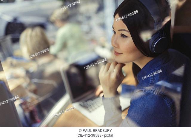 Pensive young woman at laptop listening to music with headphones at cafe window