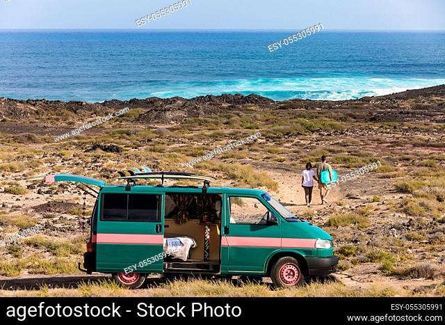 View of couple with surfboard walking on coastline with parked camping van on background of ocean, Spain