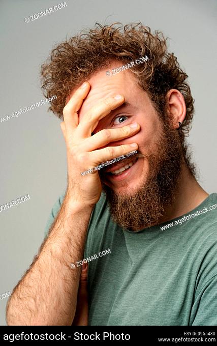 Shy handsome young man covered his face with hand pry with his eye, curly hair in olive t-shirt isolated on white background