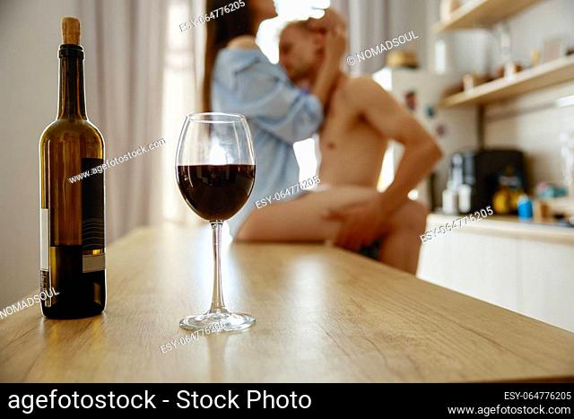 Intimacy foreplay of loving kissing couple on kitchen table at home. Focus on wineglass and bottle
