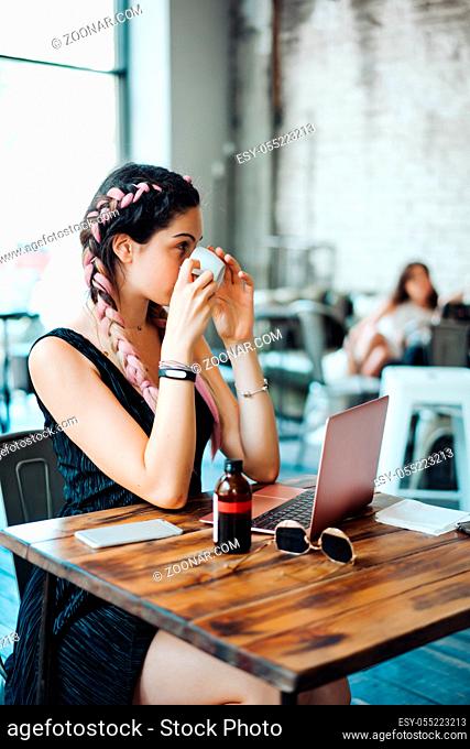 Young woman sitting in coffee shop at wooden table. On table is laptop, drinking coffee
