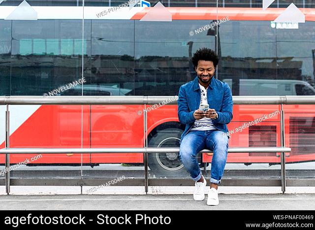 Smiling stylish mid adult man using mobile phone while sitting at bus stop