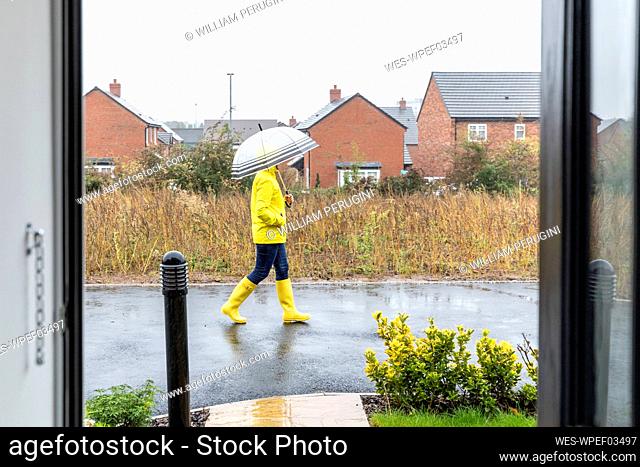 Mid adult woman with hands in pockets holding umbrella waking on street during rainy season