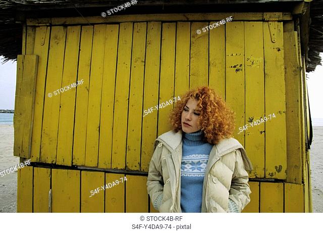 Young Woman with red curly Hair leaning against a yellow wooden Wall - Winterly Clothing - Season - Facial Expression