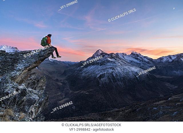 An hiker on a rock in Viola valley with a panoramic view to the Alps at sunset. Valdidentro, Valtellina, Lombardy, Italy