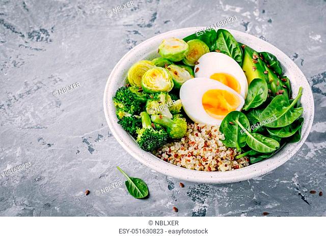 Healthy green vegetarian buddha bowl lunch with eggs, quinoa, spinach, avocado, grilled brussels sprouts and broccoli with flax seeds on dark gray background