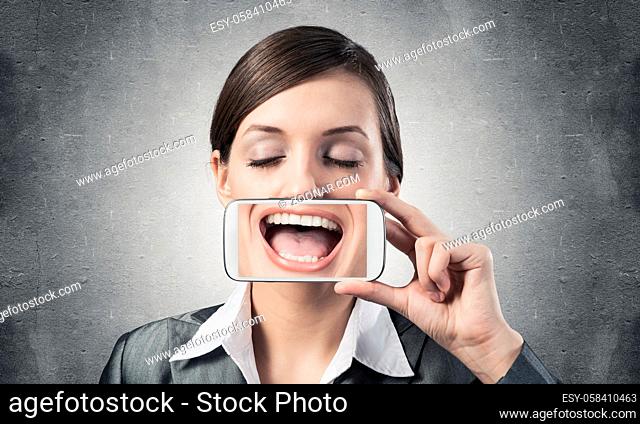 Portrait of young woman covering her mouth with smartphone. Businesswoman showing mobile phone with smile on screen. Corporate businessperson on grey wall...