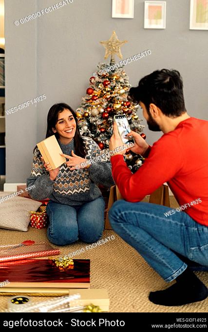 Man photographing girlfriend holding gift box sitting in front of Christmas tree
