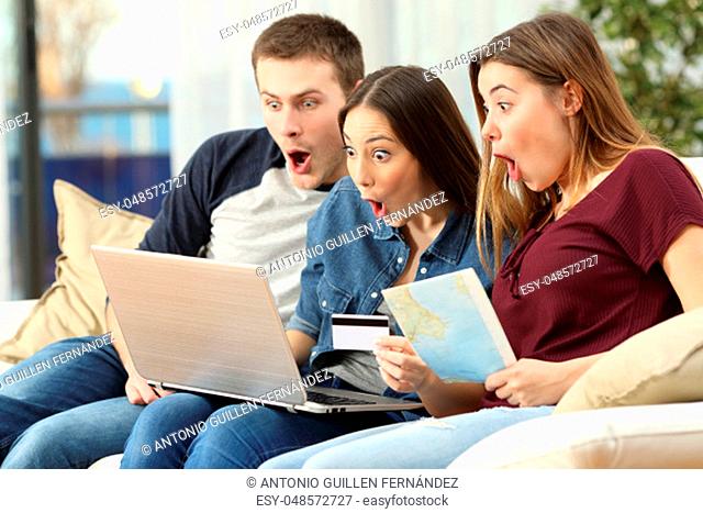 Three amazed friends finding trip offers on line with a laptop sitting on a couch in the living room in a house interior