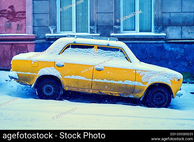 Old dirty yellow car covered with snow parked on a snowy sloping street in winter city