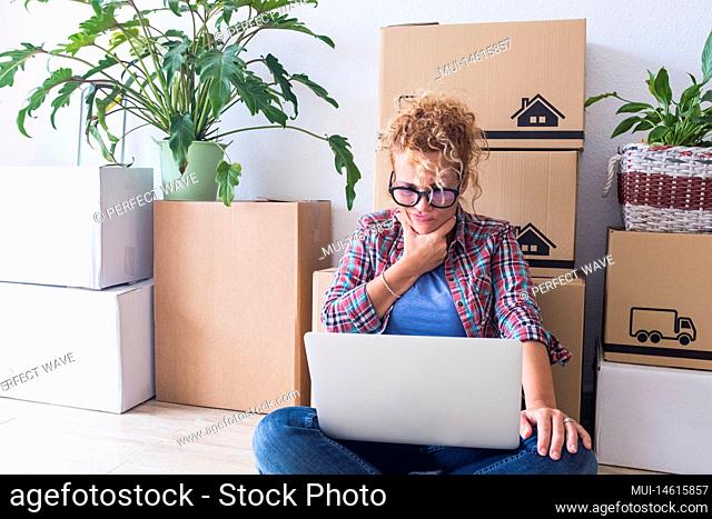 close up and portrait of young woman at home in a new house with boxes and packs in her back - adult woman using her laptop but not understanding what is in it