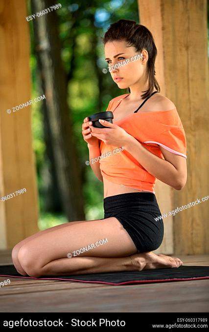 Brunette girl sits sideways on her heels on the black yoga mat on the wooden terrace on the nature background. Her head is partially turned to the left