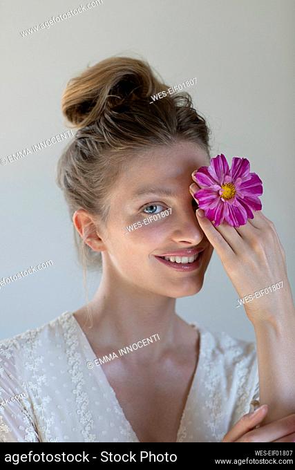 Smiling young woman covering eye with purple flower in studio
