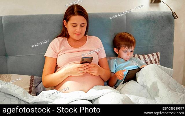 Little boy with pregnant mother in pajamas browsing internet and playing games on smartphone and tablet computer while lying under blanket at night