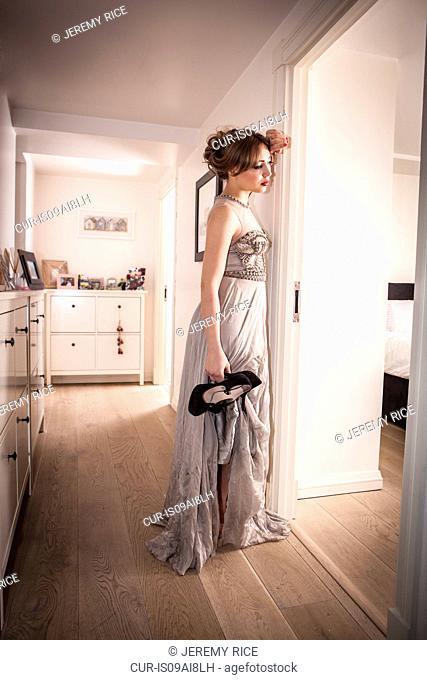 Tired young woman in evening gown, holding shoes in hallway