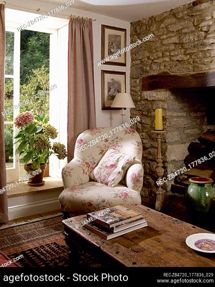 Country cottage sitting room detail with an open window, wooden coffee table, and a floral upholstered armchair next to a stone inglenook fireplace