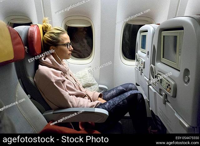 Sporty casual young blonde caucasian lady watching movie while traveling by airplane by the window. Commercial transportation by planes