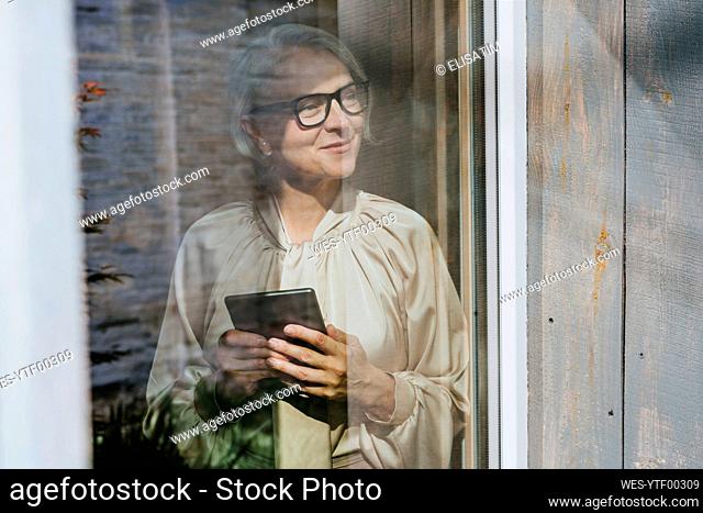Thoughtful smiling mature businesswoman holding tablet PC seen through glass window