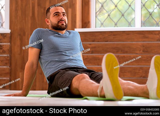 Relaxing sitting leaned back on his arms young happy fitness guy doing stretching exercises at home. Amateur sports man in workout activity sitting on pad and...