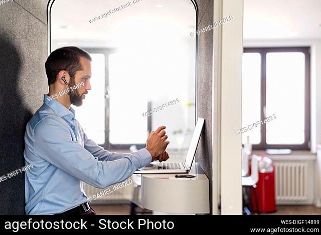 Businessman attending meeting on laptop while sitting in telephone booth at office