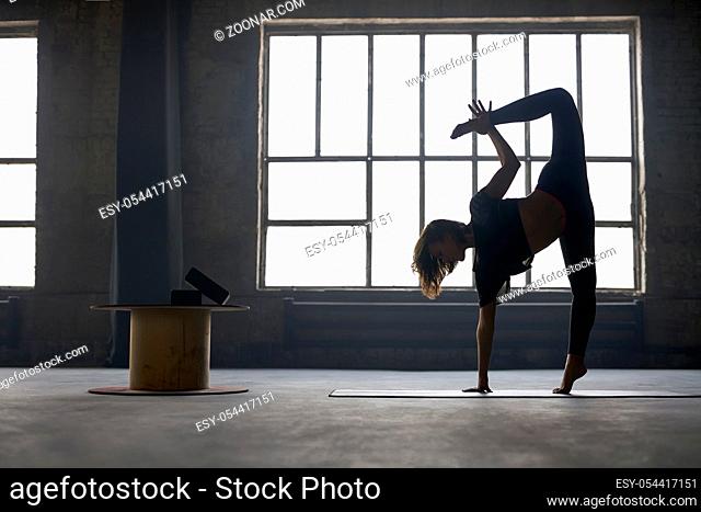 Beautiful girl in a black sportswear engaged in yoga on the window background in a loft style hall. She stands on the right leg on the mat and does a tilt