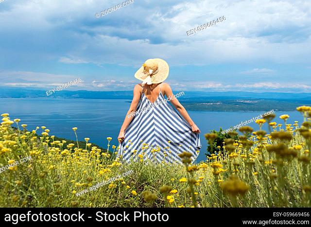 Rear view of young woman wearing striped summer dress and straw hat standing in super bloom of wildflowers, relaxing while enjoing beautiful view of Adriatic...