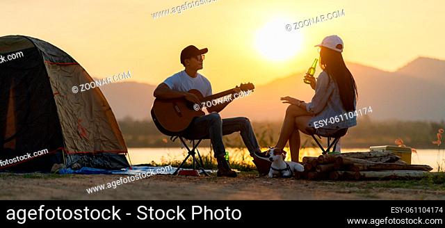 Panoramic Young adult Asian couple playing guitar and drinking beer beside their tent campsite while sunset. Camping trip with dog outdoor activity campsite...