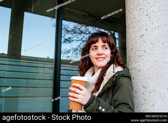 Smiling young woman holding reusable cup while looking away against column