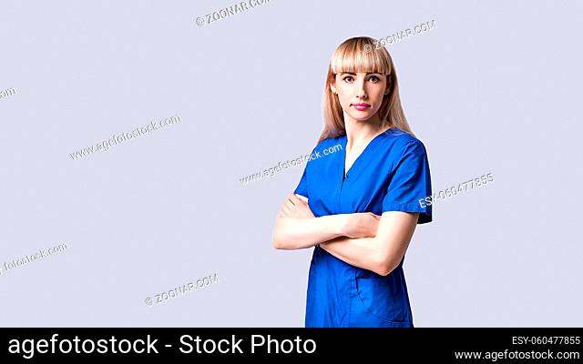 Confident young blonde woman doctor standing arms crossed. Intern in blue medical clothes posing on grey background