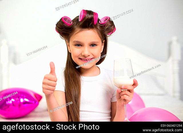 Lovely girl with hair curlers showing thumb up. Cute girlie showing thumb up and holding wineglass full of milk in the other hand in white bedroom