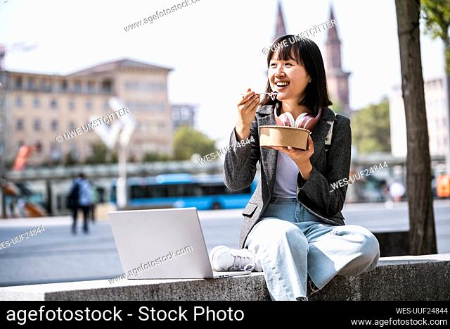 Businesswoman with laptop eating food on retaining wall
