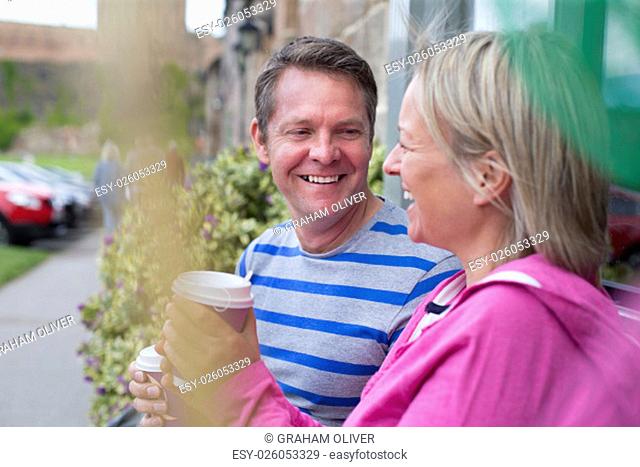 Mature couple laughing as they sit outside and drink hot drinks out of takeaway cups. They are wearing casual clothing and look very happy