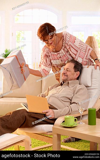 Senior couple celebrating birthday. Wife giving present to her husband, resting in armchair