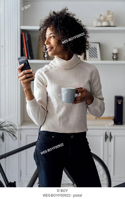 Mid adult freelancer standing in her home office, using smartphone, holding coffee cup