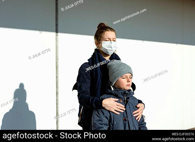 Woman wearing mask standing with son in front of a white wall, eyes closed
