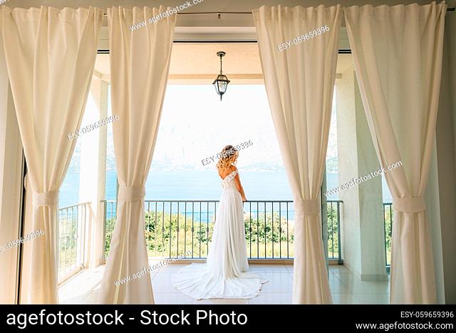 A sophisticated bride in an elegant wedding dress stands on a wide balcony with a picturesque view of the bay . High quality photo