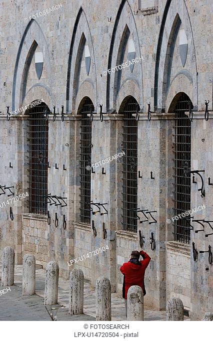 rear view of tourist in red coat, standing beside Palazzo Pubblico in Siena, photographing series of arches and windows of facade
