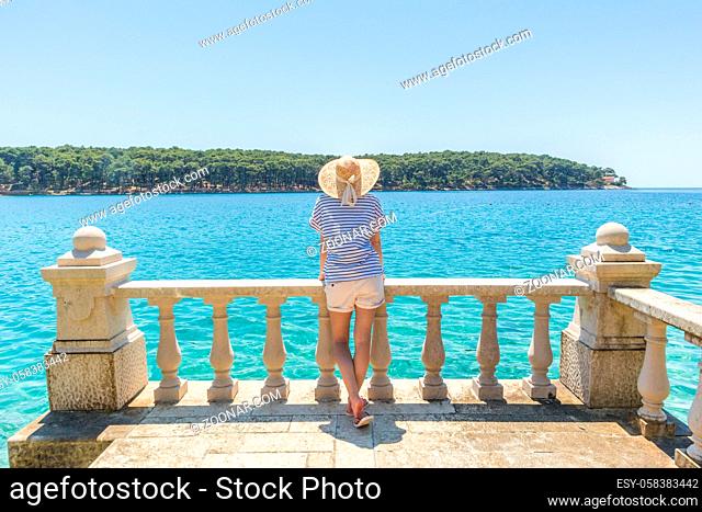 Rear view of woman wearing straw summer hat , leaning against elegant old stone fence of coastal villa, relaxing while looking at blue Adriatic sea