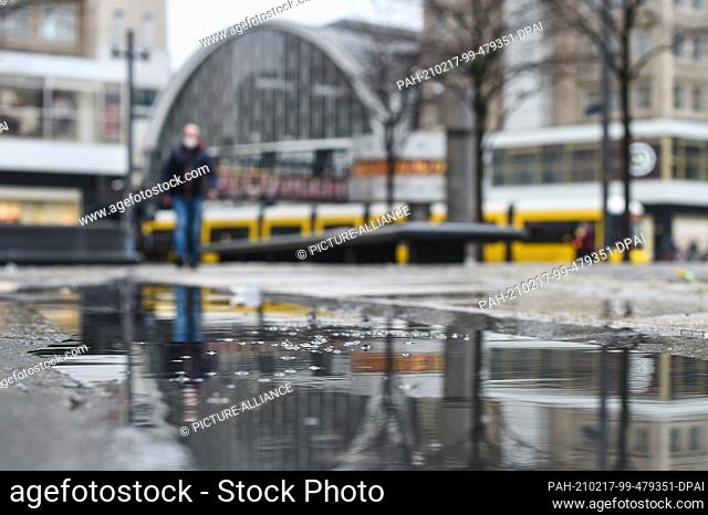 17 February 2021, Berlin: In a puddle of rain on Alexanderplatz, the S-Bahn station, a tram and a person with mouth-nose covering to see