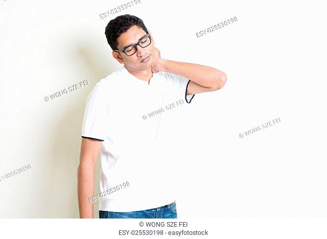 Portrait of Indian guy neck pain, massaging with tired face expression. Asian man standing on plain background with shadow and copy space