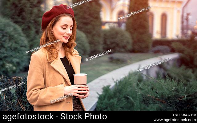 Beautiful young woman holding a mug with coffee standing outdoors. Portrait of stylish young woman wearing autumn coat and red beret outdoors