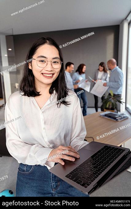 Portrait of smiling young businesswoman using laptop with colleagues in background in office
