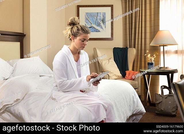 Young Caucasian woman wearing bathrobe in a hotel room, using tablet device sitting on edge of bed