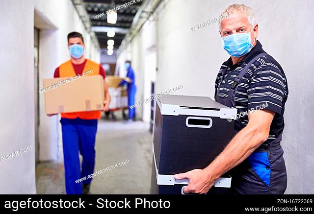Älterer Arbeiter mit Mundschutz wegen Covid-19 trägt Pakete ins Lager einer Fabrik