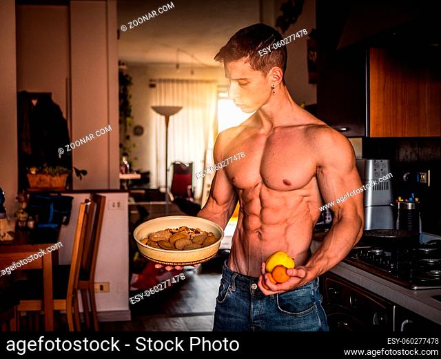 Muscular shirtless young man deciding between healthy fruit and unhealthy cookies at home in kitchen