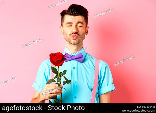 Cute and funny man waiting for kiss from lover on Valentines day, holding beautiful red rose for girlfriend, standing over pink background