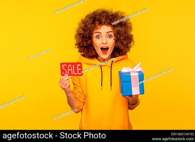 Wow, bonuses on Black Friday shopping. Portrait of surprised overjoyed curly-haired woman in urban style hoodie showing Sale inscription and gift box