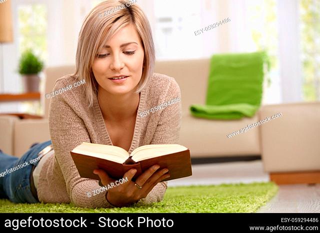 Young blonde woman relaxing on floor at home reading book. Copyspace on right
